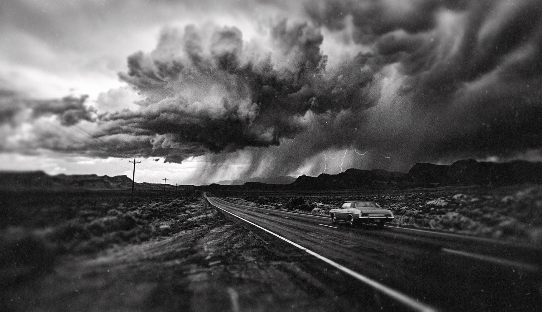 Black-and-white photograph: a lone car on a desert highway driving toward a vast storm cloud with lightning.